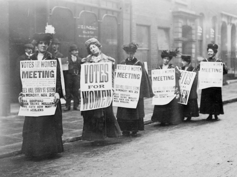 Women in full-length dresses march with signs reading “VOTES FOR WOMEN” in a historical photo from the Suffrage Era.