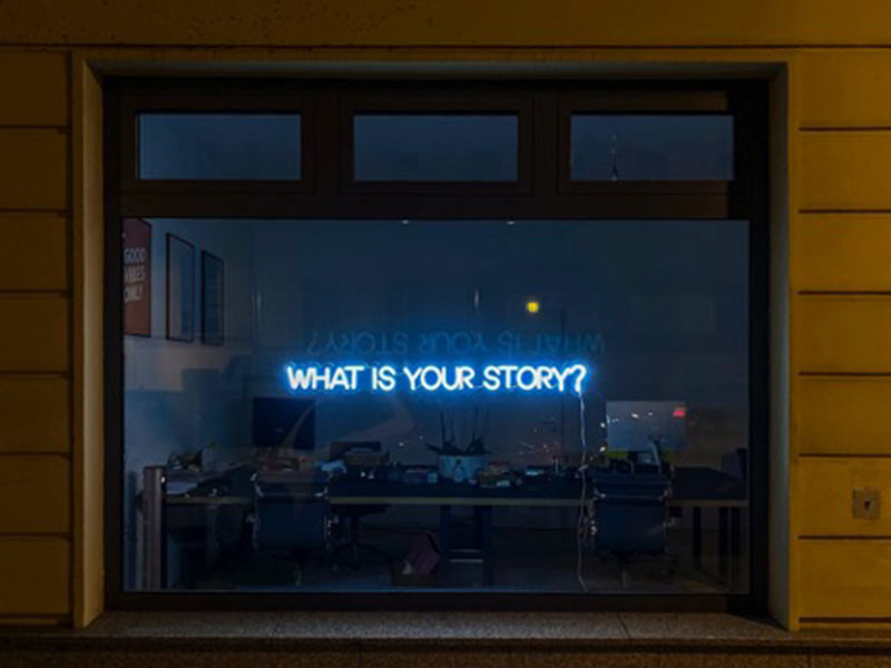 After dark, a neon sign hanging in a street-level window glows with the words “WHAT IS YOUR STORY?”