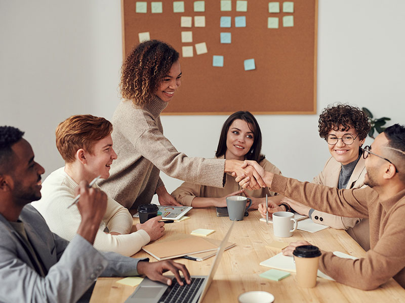 A diverse group people sitting around a table for a meeting, with colleagues shaking each other's hands.