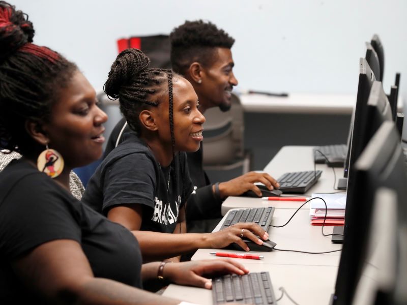 Several CCAC students smile as they work at a row of computers.