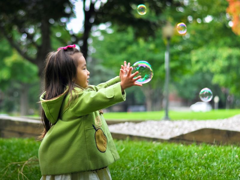 A little girl smiles as she chases bubbles through a grassy park.
