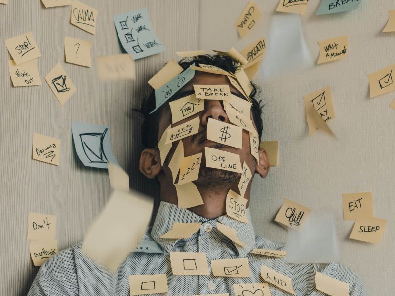 A man in a button-down shirt is covered with sticky notes indicating his stressful to-do list.