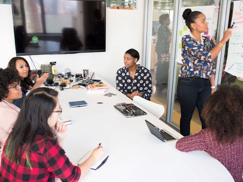 Half a dozen women talk around a conference table while one takes notes on a flipchart.