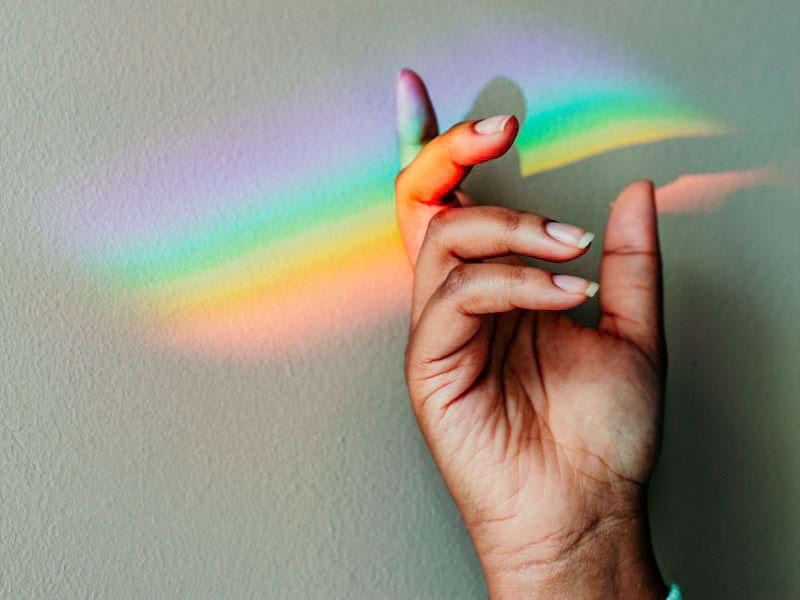 Rainbow light from a prism illuminates someone's hand against a white wall.